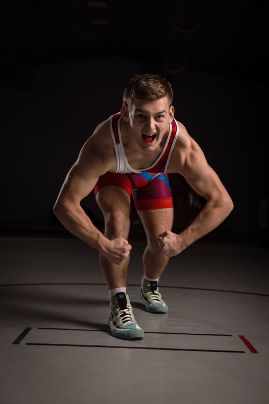 Wrestler in a red, white, and blue singlet crouching and shouting toward the camera in a dark gym