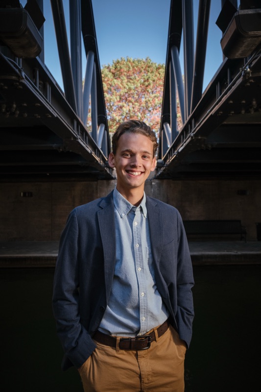 Smiling young man in a navy blazer standing under a steel bridge