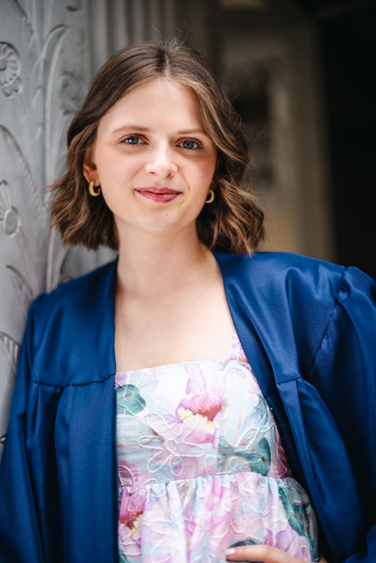 Smiling young woman in a navy graduation gown over a floral dress, leaning on a carved stone doorway