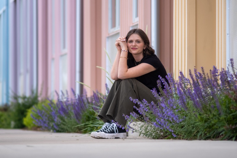 Young woman in a black T-shirt and high-top sneakers sitting among purple flowers beside a pastel-striped wall